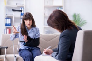 A young woman holding a crutch with her arm in a sling is talking to a woman wearing a blazer.