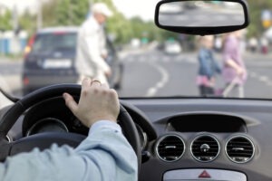 A driver has one hand on the wheel and is approaching pedestrians in a crosswalk