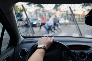 A driver looking through a rainy windshield approaches a group of pedestrians in a crosswalk.