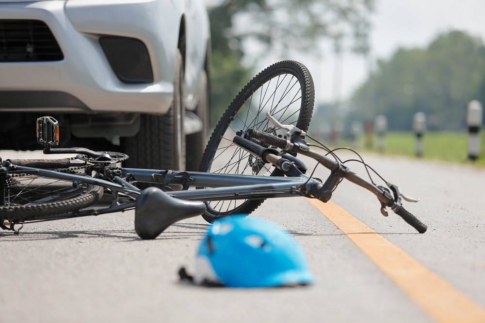Bicycle lying on the road in front of a silver car, with a blue helmet on the pavement