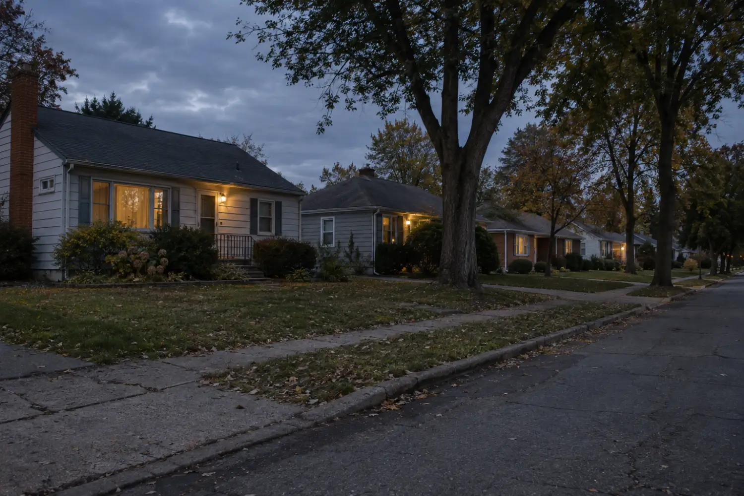 Quiet residential street at dusk with porch lights on along a row of single-story houses
