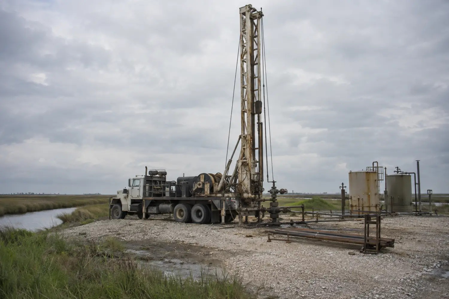 Truck-mounted workover rig raised over a well on a flat coastal lease with marsh grass nearby