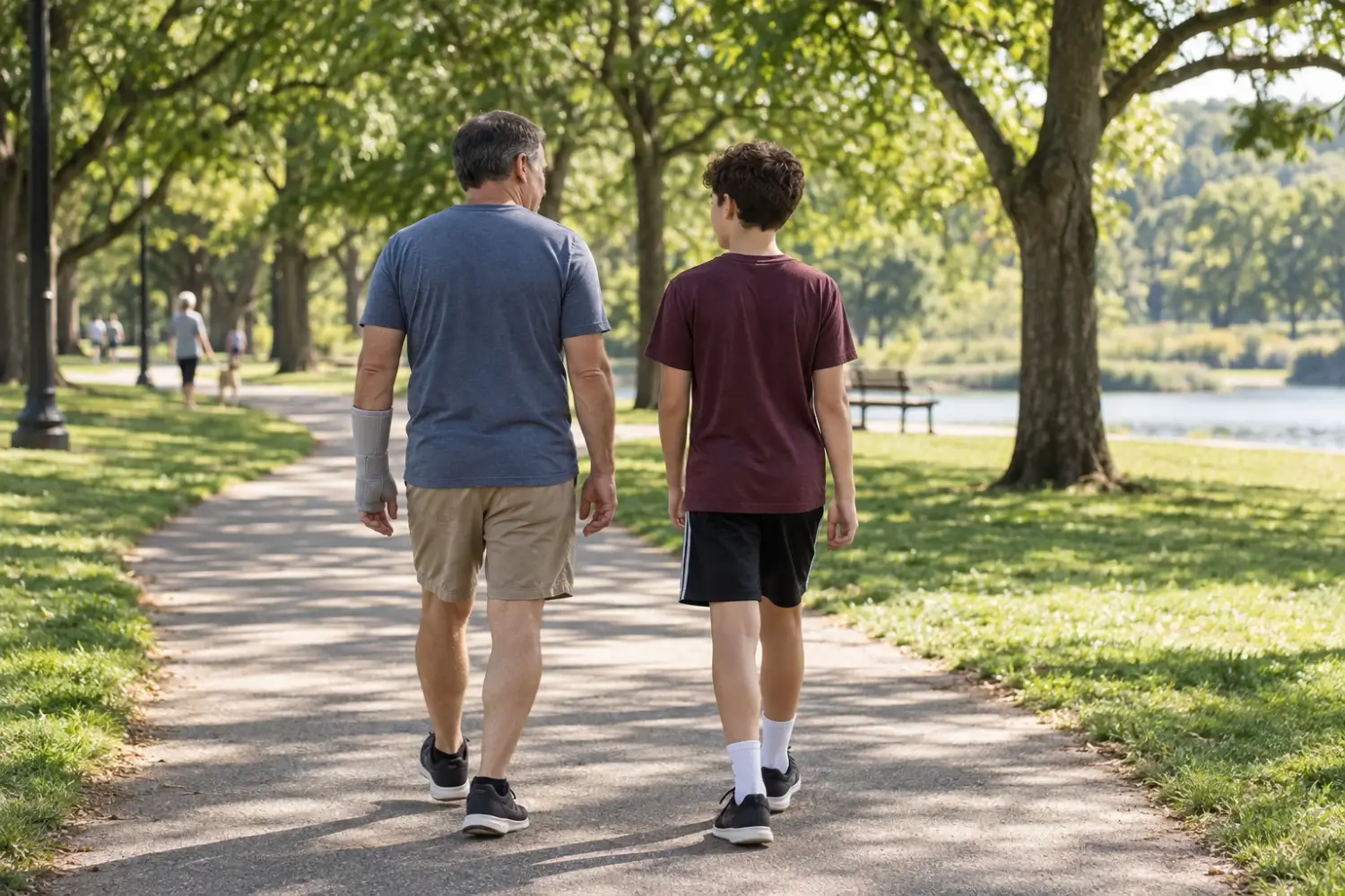 Adult wearing a forearm brace walks beside a teenager on a shaded park path, both seen from behind