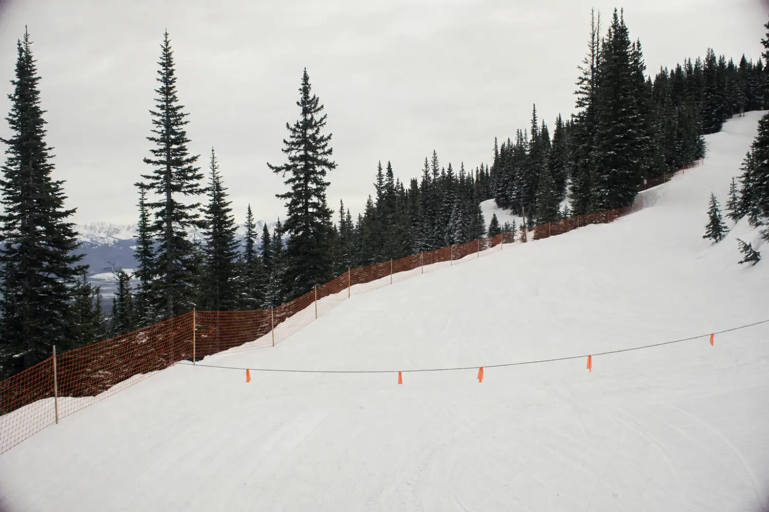 Groomed ski run with orange safety fencing and a rope line with orange flags across a closed section