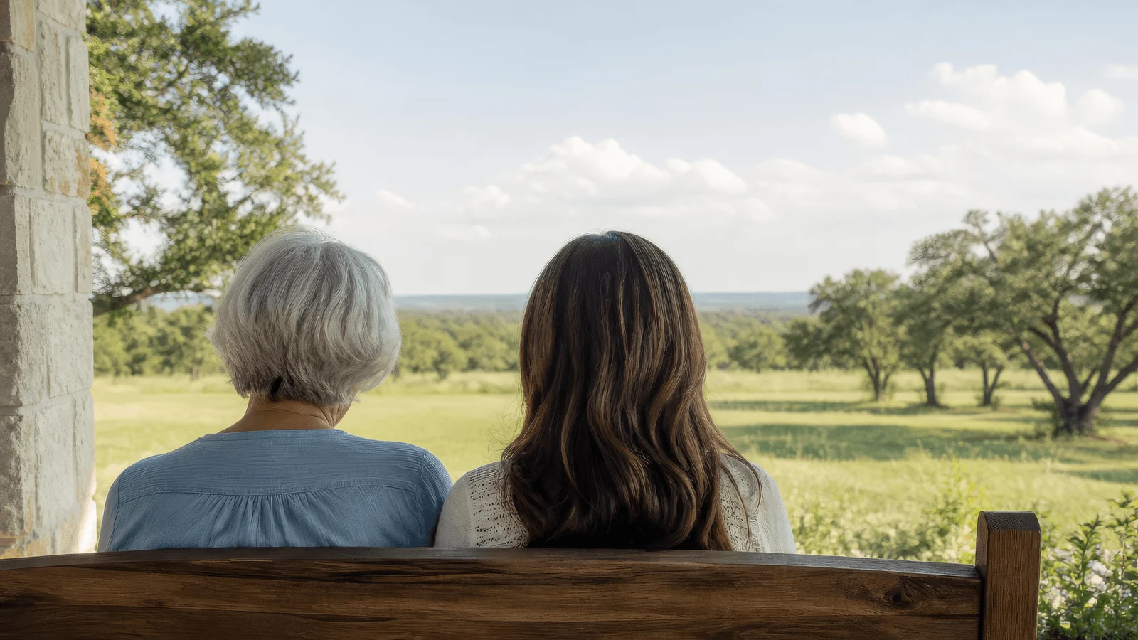 An older woman and her adult daughter sit side by side on a porch bench, looking out over a Texas field.