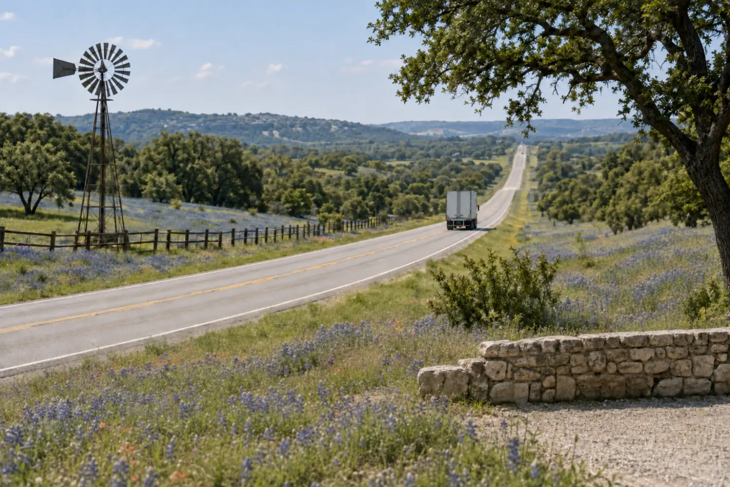 Tractor-trailer on a Texas Hill Country highway lined with bluebonnets, with a windmill and a stone wall by the road